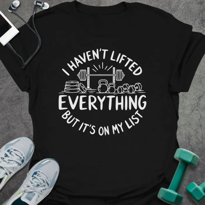Black t-shirt printed with "I HAVEN'T LIFTED EVERYTHING BUT IT'S ON MY LIST" in white, with a barbell, weight plates, kettlebells and dumbbells