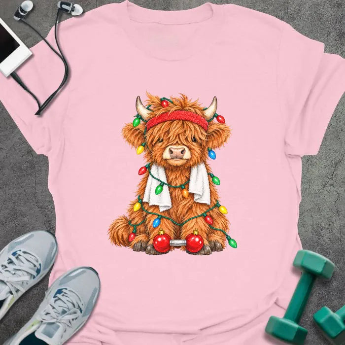 Light pink t-shirt printed with a fluffy highland cow in a red headband and towel, tangled in Christmas lights beside a barbell with ornament plates