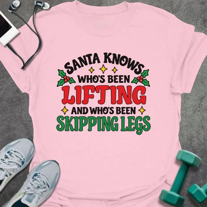 Light pink t-shirt printed with "SANTA KNOWS WHO'S BEEN LIFTING AND WHO'S BEEN SKIPPING LEGS" in black, red and green, with holly and gold stars