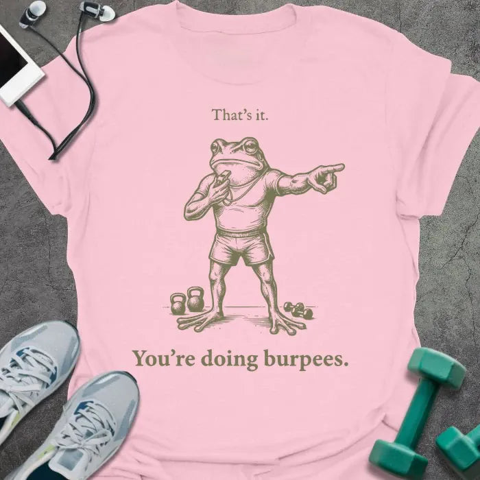 Light pink t-shirt printed with "That's it. You're doing burpees." in tan, with an illustration of a frog character in sportswear pointing