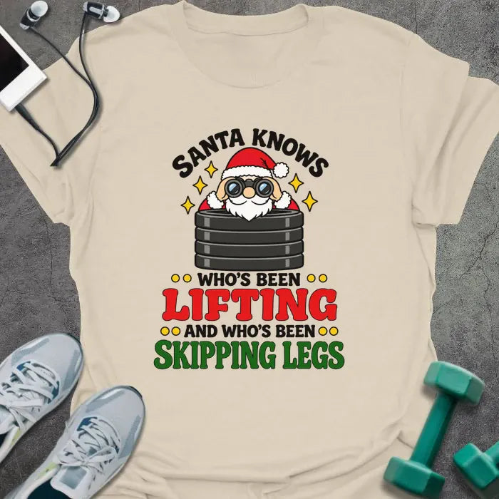 Natural t-shirt printed with "SANTA KNOWS WHO'S BEEN LIFTING AND WHO'S BEEN SKIPPING LEGS" in black, red and green, with Santa peering through binoculars over a stack of weight plates