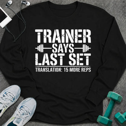 Black long sleeve tee printed with "Trainer Says Last Set. Translation: 15 More Reps" in white, with barbell accents