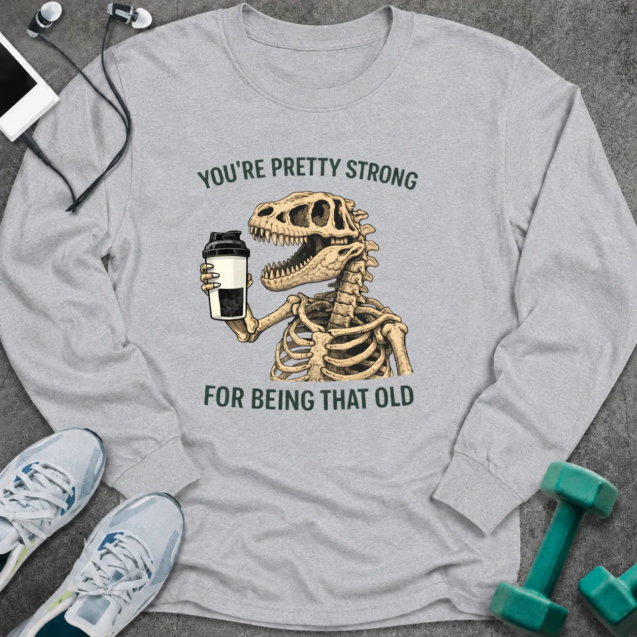 Ash long sleeve tee printed with "You're Pretty Strong For Being That Old" in green, with a dinosaur skeleton holding a shaker cup
