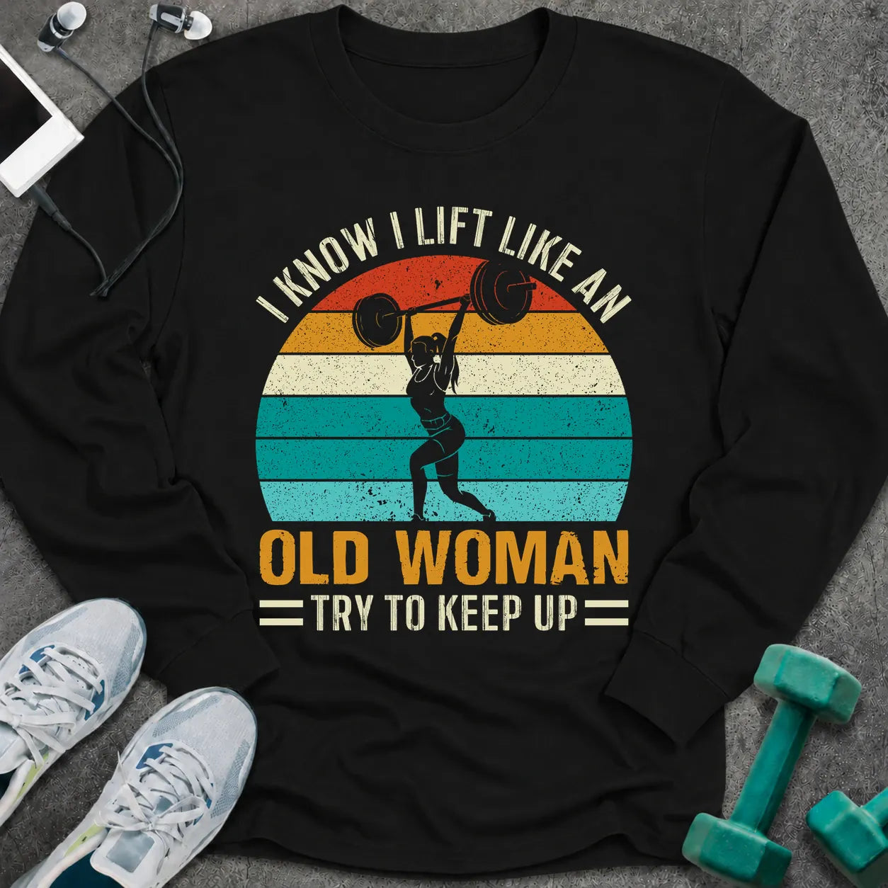 Black long sleeve tee printed with "I Know I Lift Like An Old Woman, Try To Keep Up" in white and gold, with a woman pressing a barbell over a retro striped sunset
