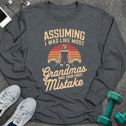 Dark heather long sleeve tee printed with "Assuming I Was Like Most Grandmas Was Your First Mistake" in cream and gold, with a woman deadlifting inside a retro sunburst