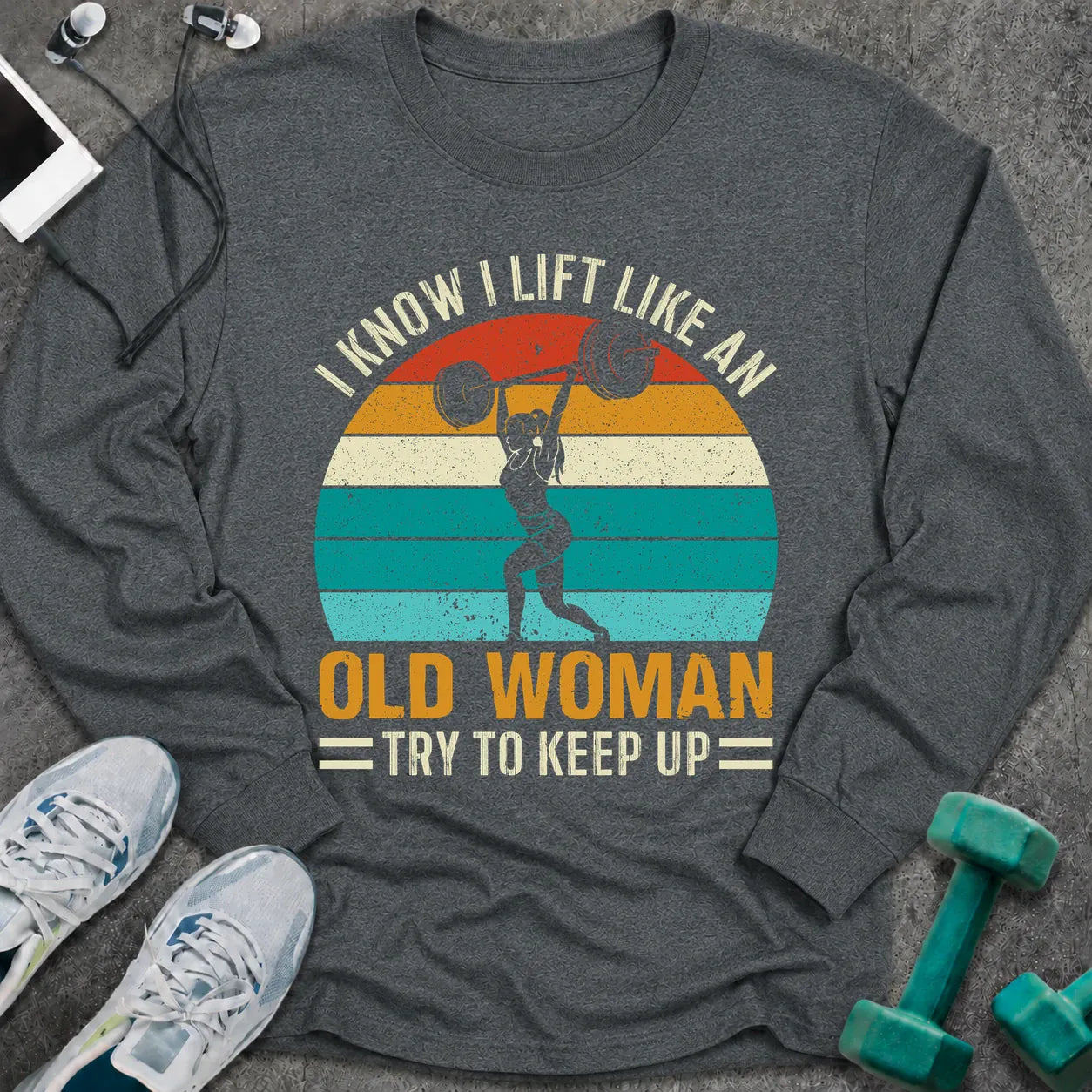 Dark heather long sleeve tee printed with "I Know I Lift Like An Old Woman, Try To Keep Up" in white and gold, with a woman pressing a barbell over a retro striped sunset