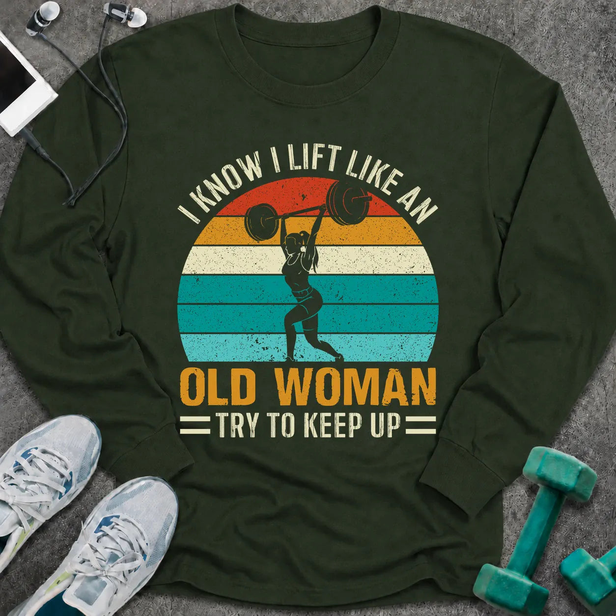 Forest green long sleeve tee printed with "I Know I Lift Like An Old Woman, Try To Keep Up" in white and gold, with a woman pressing a barbell over a retro striped sunset