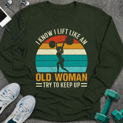 Forest green long sleeve tee printed with "I Know I Lift Like An Old Woman, Try To Keep Up" in white and gold, with a woman pressing a barbell over a retro striped sunset
