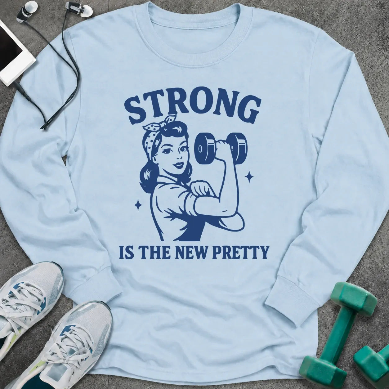 Light blue long sleeve tee printed with "Strong Is The New Pretty" in navy, with a retro woman flexing with a dumbbell
