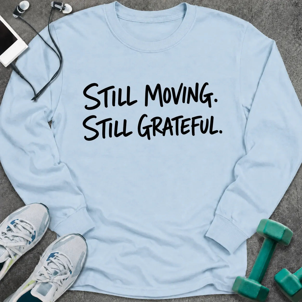Light blue long sleeve tee printed with "Still Moving. Still Grateful." in black handwritten lettering