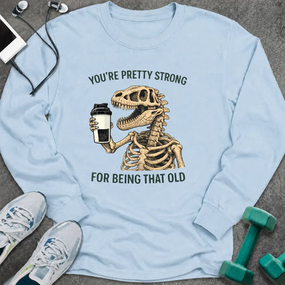 Light blue long sleeve tee printed with "You're Pretty Strong For Being That Old" in green, with a dinosaur skeleton holding a shaker cup