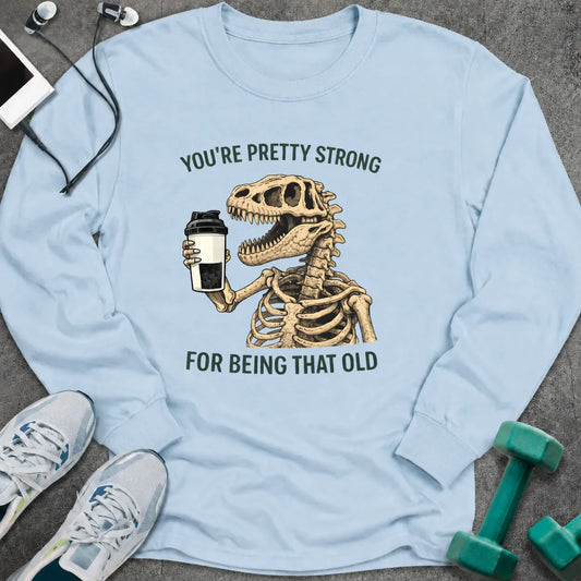 Light blue long sleeve tee printed with "You're Pretty Strong For Being That Old" in green, with a dinosaur skeleton holding a shaker cup