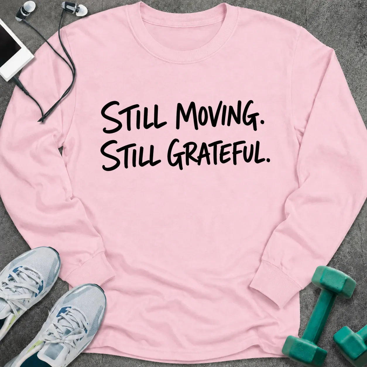 Light pink long sleeve tee printed with "Still Moving. Still Grateful." in black handwritten lettering