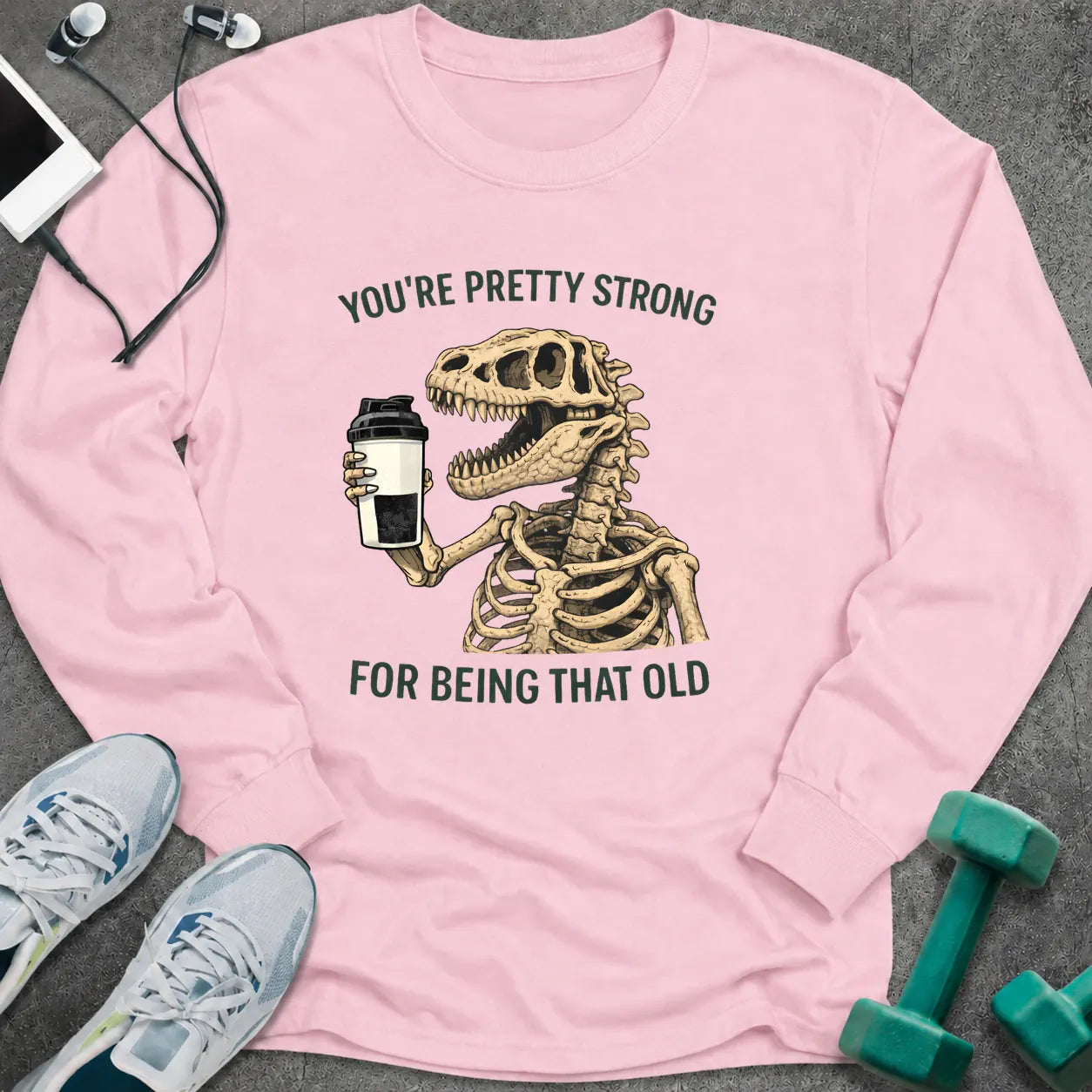 Light pink long sleeve tee printed with "You're Pretty Strong For Being That Old" in green, with a dinosaur skeleton holding a shaker cup