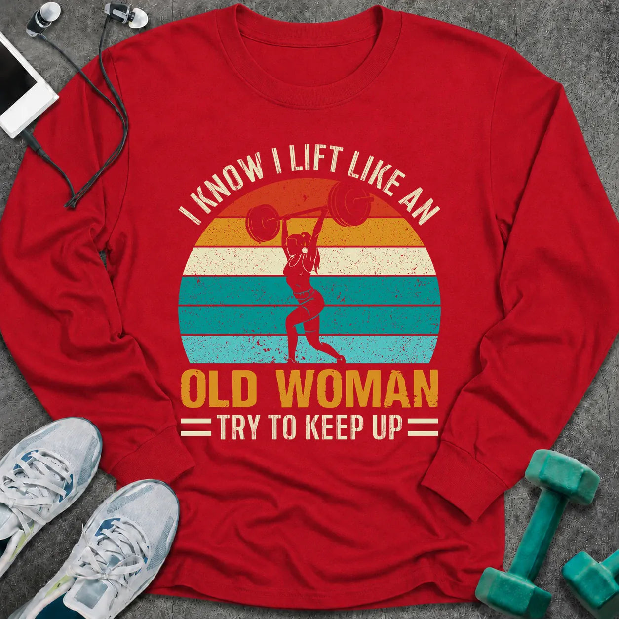 Red long sleeve tee printed with "I Know I Lift Like An Old Woman, Try To Keep Up" in white and gold, with a woman pressing a barbell over a retro striped sunset