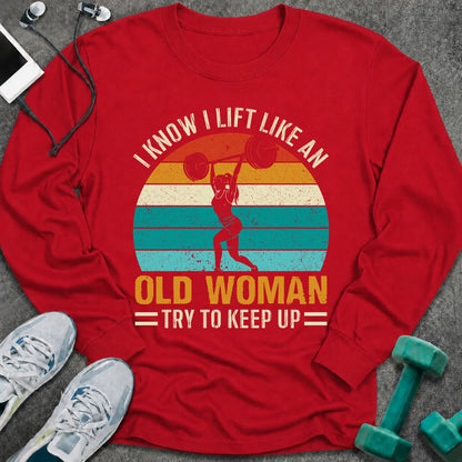 Red long sleeve tee printed with "I Know I Lift Like An Old Woman, Try To Keep Up" in white and gold, with a woman pressing a barbell over a retro striped sunset