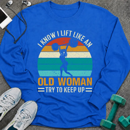 Royal long sleeve tee printed with "I Know I Lift Like An Old Woman, Try To Keep Up" in white and gold, with a woman pressing a barbell over a retro striped sunset