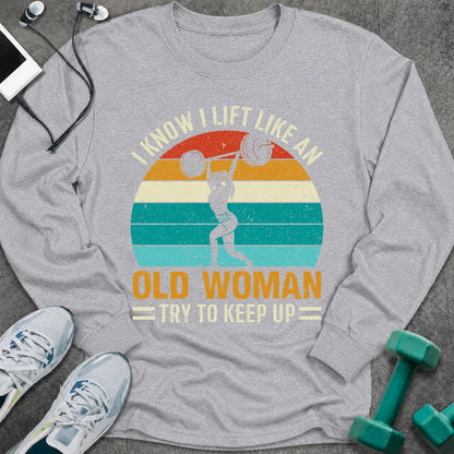 Sport grey long sleeve tee printed with "I Know I Lift Like An Old Woman, Try To Keep Up" in white and gold, with a woman pressing a barbell over a retro striped sunset
