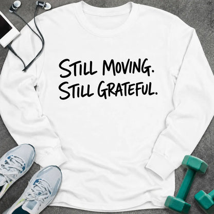 White long sleeve tee printed with "Still Moving. Still Grateful." in black handwritten lettering