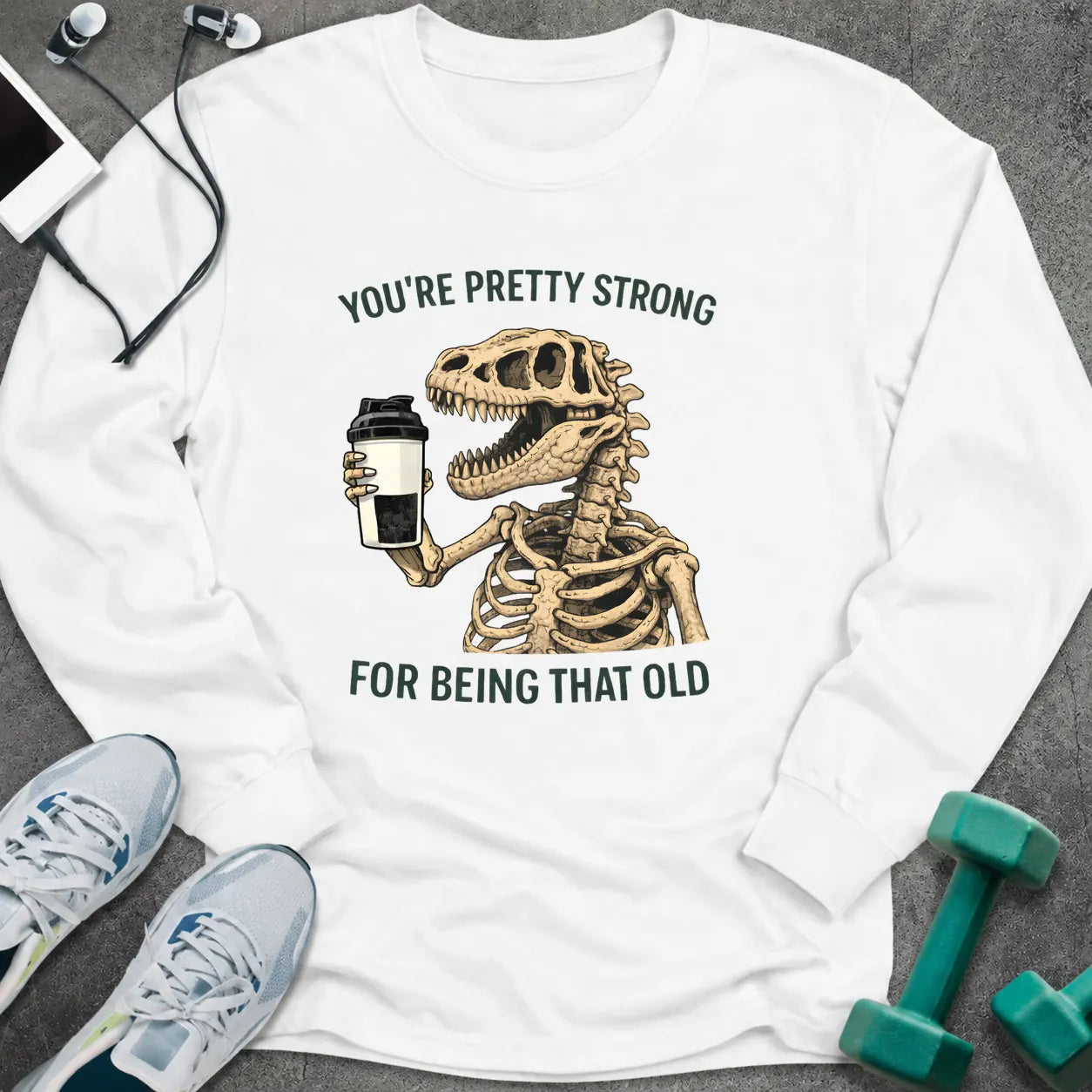 White long sleeve tee printed with "You're Pretty Strong For Being That Old" in green, with a dinosaur skeleton holding a shaker cup