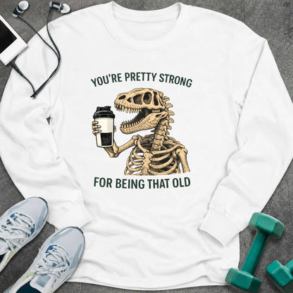 White long sleeve tee printed with "You're Pretty Strong For Being That Old" in green, with a dinosaur skeleton holding a shaker cup