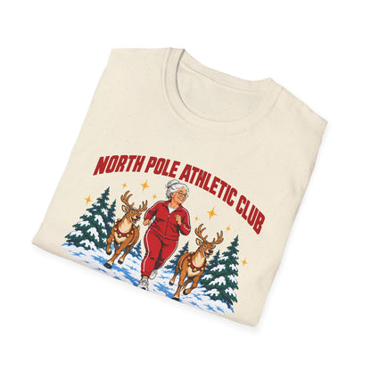 Folded natural t-shirt printed with "NORTH POLE ATHLETIC CLUB" in red and "Winter Training Season" in green script, with a white-haired woman in a red tracksuit jogging between two reindeer through snowy pine trees