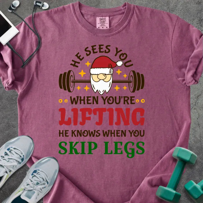Berry t-shirt printed with "He Sees You When You're Lifting He Knows When You Skip Legs" in multicolor, with Santa holding a barbell