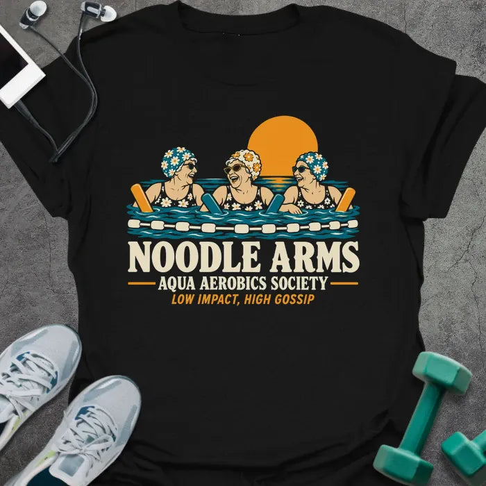 Black t-shirt printed with "NOODLE ARMS, AQUA AEROBICS SOCIETY, LOW IMPACT, HIGH GOSSIP" in cream and orange, with three older women in flowered swim caps and sunglasses laughing in a pool with pool noodles at sunset