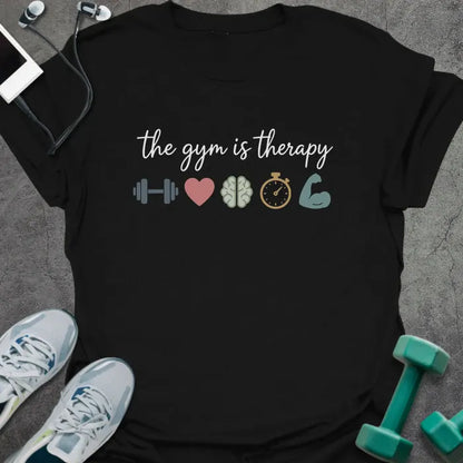 Black t-shirt printed with "the gym is therapy" in gray, pink, gray, gold, and green, with a dumbbell, heart, brain, stopwatch, and bicep emoji