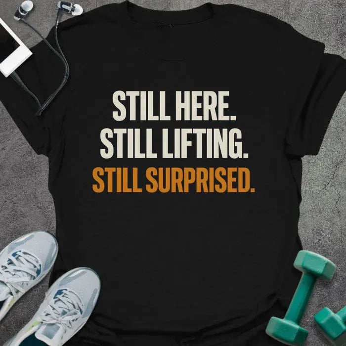 Black t-shirt printed with "Still Here. Still Lifting. Still Surprised." in white and orange