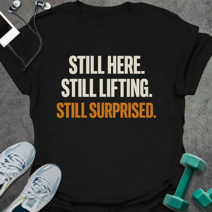 Black t-shirt printed with "Still Here. Still Lifting. Still Surprised." in white and orange