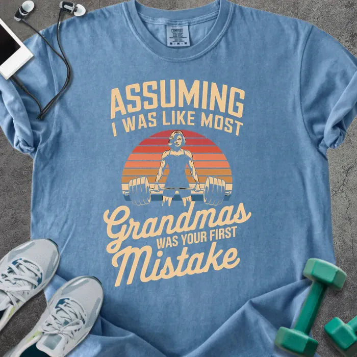 Blue jean t-shirt printed with "Assuming I Was Like Most Grandmas Was Your First Mistake" in white and gold, with a grandma lifting dumbbells graphic