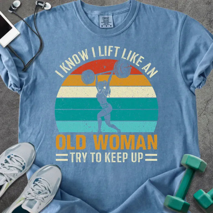 Blue jean t-shirt printed with "I Know I Lift Like an Old Woman Try to Keep Up" in orange and multicolor, with a lifting silhouette and rainbow sunset