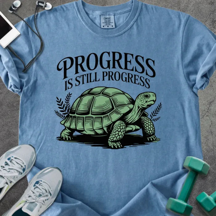 Blue jean t-shirt printed with "Progress is Still Progress" in green, with a turtle illustration and decorative leaves