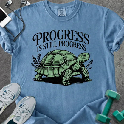 Blue jean t-shirt printed with "Progress is Still Progress" in green, with a turtle illustration and decorative leaves