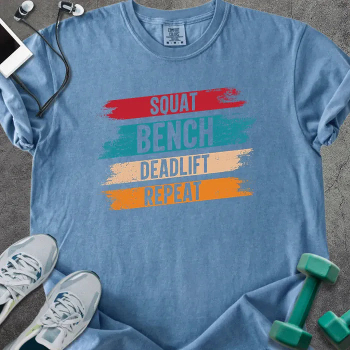 Blue jean t-shirt printed with "Squat Bench Deadlift Repeat" in red, teal, tan, orange, with red, teal, tan, and orange paint-stroke bands behind the words