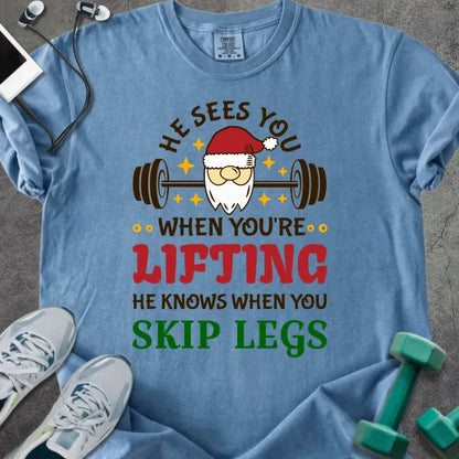 Blue jean t-shirt printed with "He Sees You When You're Lifting He Knows When You Skip Legs" in multicolor, with Santa holding a barbell