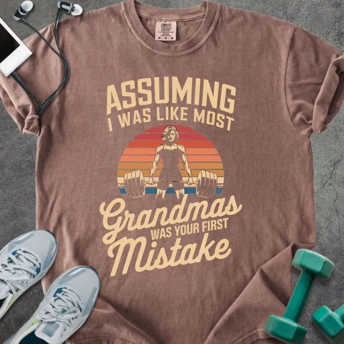Espresso t-shirt printed with "Assuming I Was Like Most Grandmas Was Your First Mistake" in white and gold, with a grandma lifting dumbbells graphic