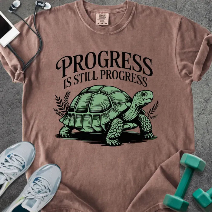 Espresso t-shirt printed with "Progress is Still Progress" in green, with a turtle illustration and decorative leaves