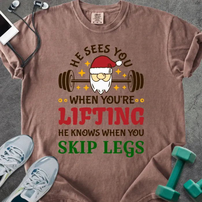 Espresso t-shirt printed with "He Sees You When You're Lifting He Knows When You Skip Legs" in multicolor, with Santa holding a barbell