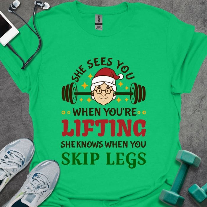 Irish green t-shirt printed with "She Sees You When You're Lifting Sheknows When You Skip Legs" in black, red, and gold, with female Santa and barbell