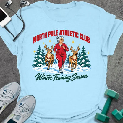 Light blue t-shirt printed with "NORTH POLE ATHLETIC CLUB" in red and "Winter Training Season" in green script, with a white-haired woman in a red tracksuit jogging between two reindeer through snowy pine trees