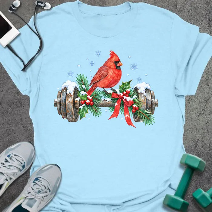 Light blue t-shirt printed with a watercolor red cardinal perched on a snowy dumbbell trimmed with holly, pine branches and a red bow, with blue snowflakes