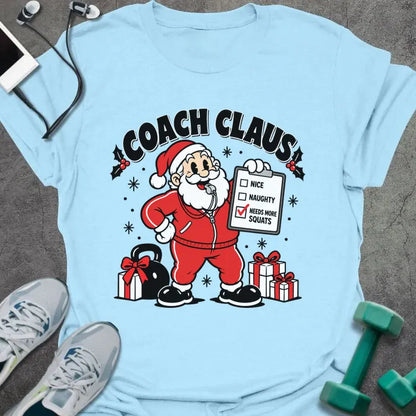 Light blue t-shirt printed with "COACH CLAUS" in black, with a cartoon Santa in a red tracksuit and whistle holding a clipboard with "NEEDS MORE SQUATS" checked, next to a kettlebell and wrapped presents