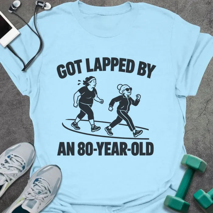 Light blue t-shirt printed with "GOT LAPPED BY AN 80-YEAR-OLD" in black, with a tired jogger being passed by an older woman in sunglasses speed walking on a track