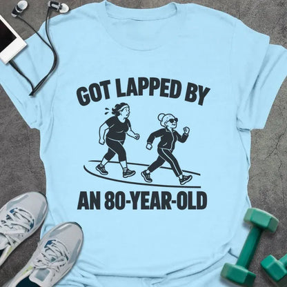 Light blue t-shirt printed with "GOT LAPPED BY AN 80-YEAR-OLD" in black, with a tired jogger being passed by an older woman in sunglasses speed walking on a track