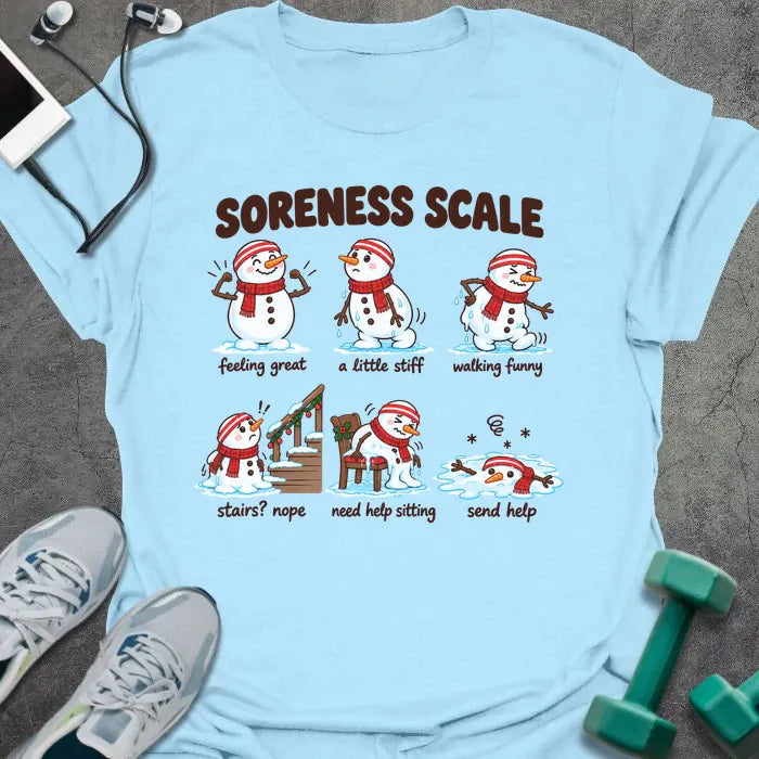 Light blue t-shirt printed with "SORENESS SCALE" in brown, with six snowmen in striped headbands and red scarves labeled feeling great, a little stiff, walking funny, stairs? nope, need help sitting, and send help