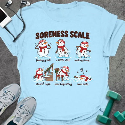Light blue t-shirt printed with "SORENESS SCALE" in brown, with six snowmen in striped headbands and red scarves labeled feeling great, a little stiff, walking funny, stairs? nope, need help sitting, and send help
