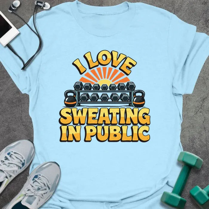 Light blue t-shirt printed with "I LOVE SWEATING IN PUBLIC" in yellow and orange, with a dumbbell rack, kettlebells and a retro sunset