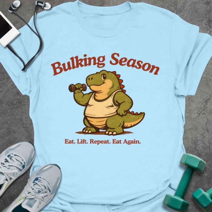 Light blue t-shirt printed with "Bulking Season Eat. Lift. Repeat. Eat Again." in brown and green, with dinosaur lifting weights graphic